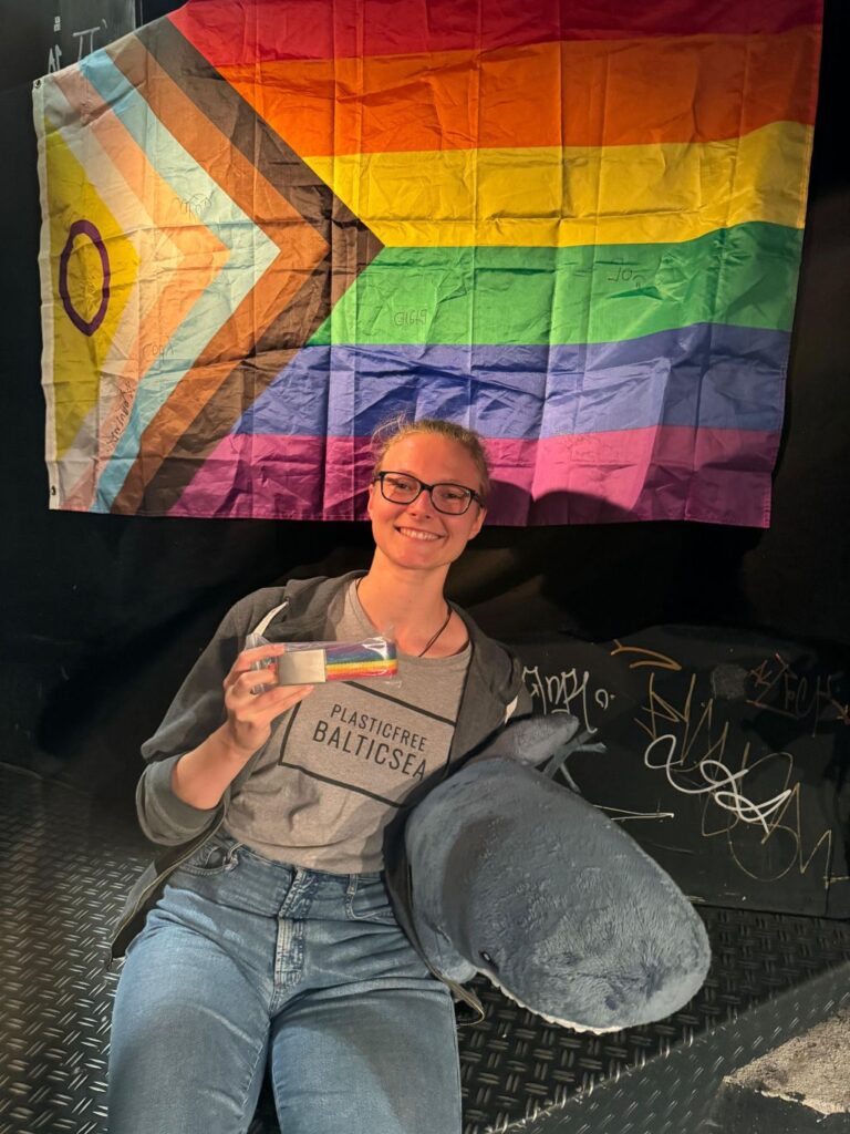 Selina Mutzbauer vor einer Regenbogenflagge. In der einen Hand hält sie einen Regenbogenfarbenen Gürtel, in der anderen einen Blahaj.