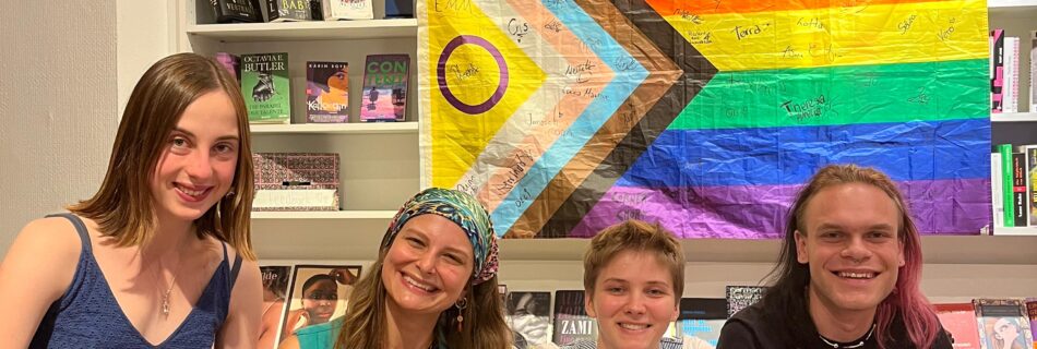 vier Personen auf einer Couch sitzend. Im Hintergrund ein Bücherregal an welchem eine Regenbogenflagge hängt.